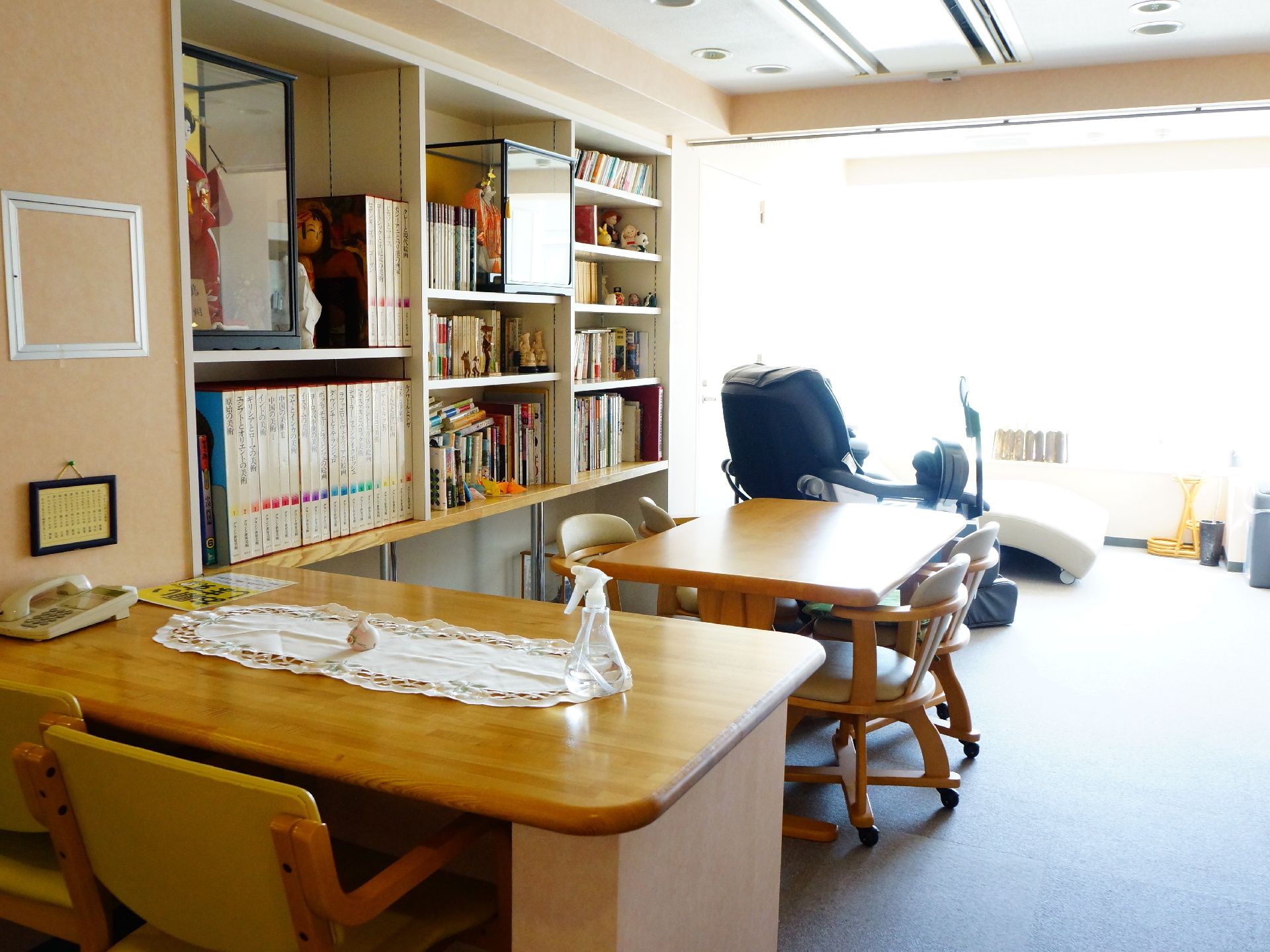 左の棚には日本人形や書物がなどが敷き詰められている。光の入る窓にはマッサージチェアが置かれ、他にも2台の机がある。