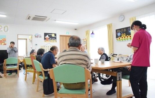 共有スペースに、男性、女性の入居者様、車椅子をご利用の方が着席しており、女性スタッフの話を聞いている。