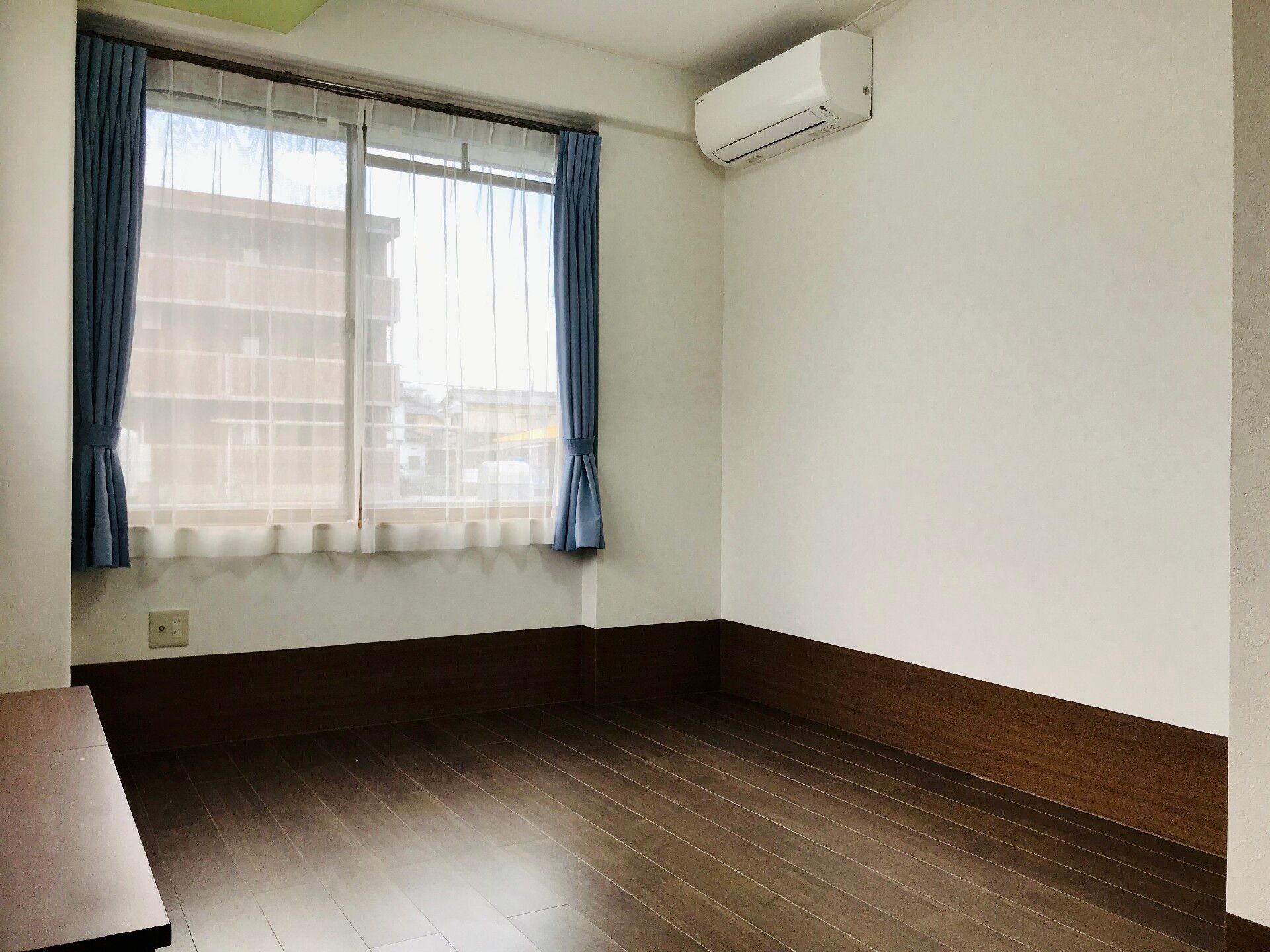 落ち着いたフローリングの床面が特徴的な居室である。奥の壁には大きめの窓が取りつけられており、青色のカーテンが留められている。