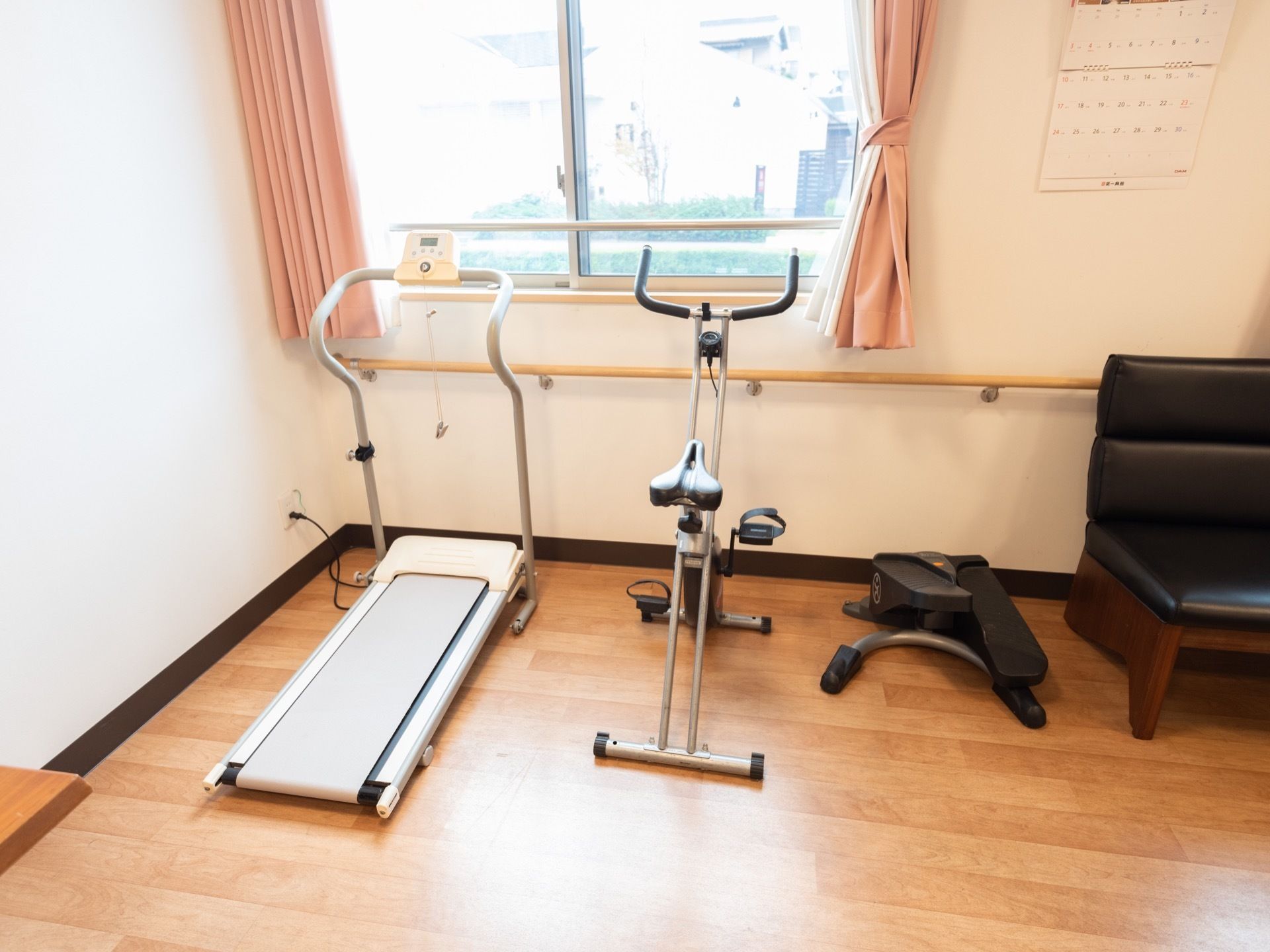 窓の下に手すりが設置されており、その手前にリハビリ器具が並べて置かれている。窓の右側にカレンダーが掛けられている。
