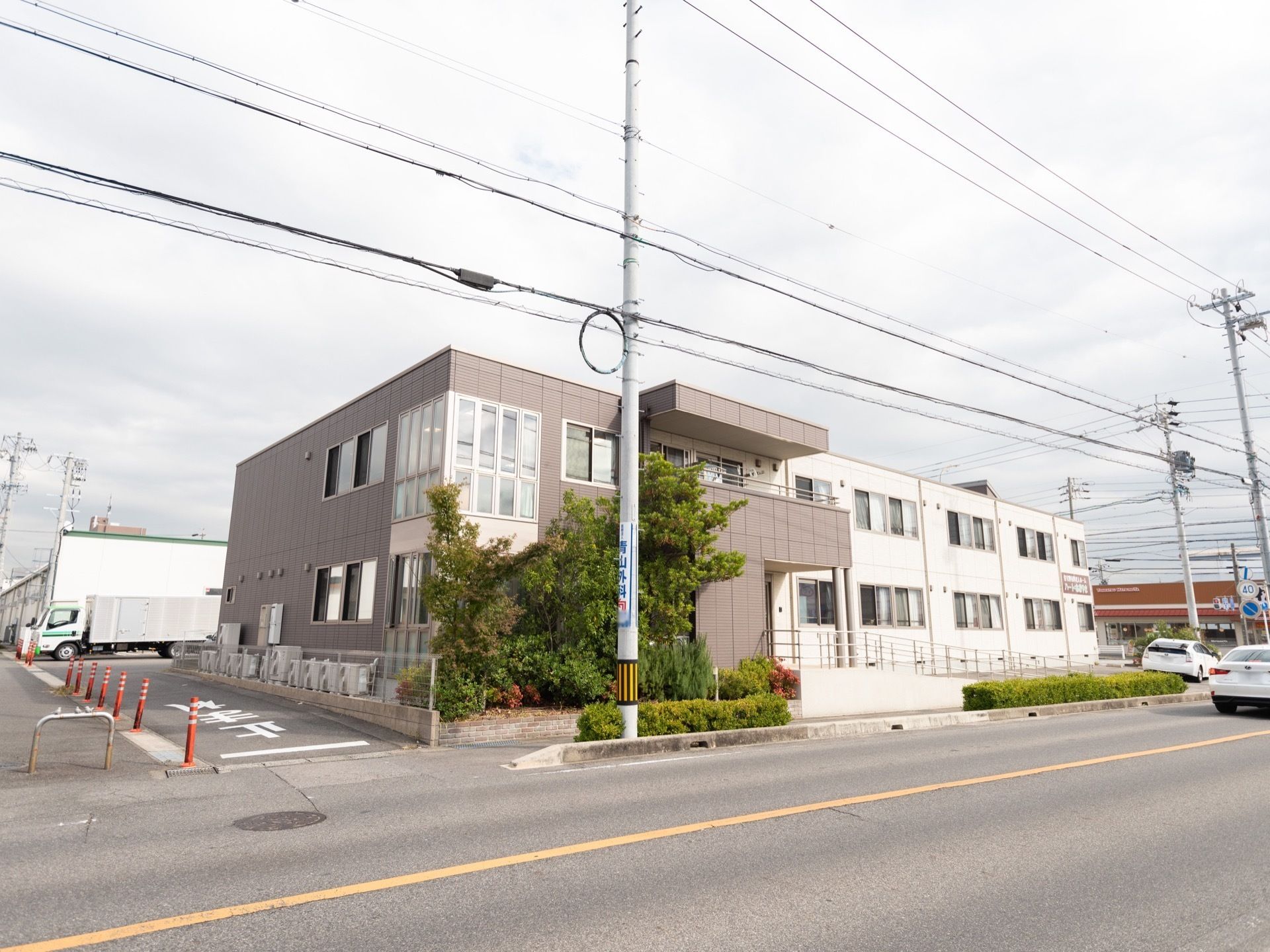 白とグレーの落ち着いた色合いの外壁の建物で、施設前に2階までの高さのある樹木がたくさん植えられている。