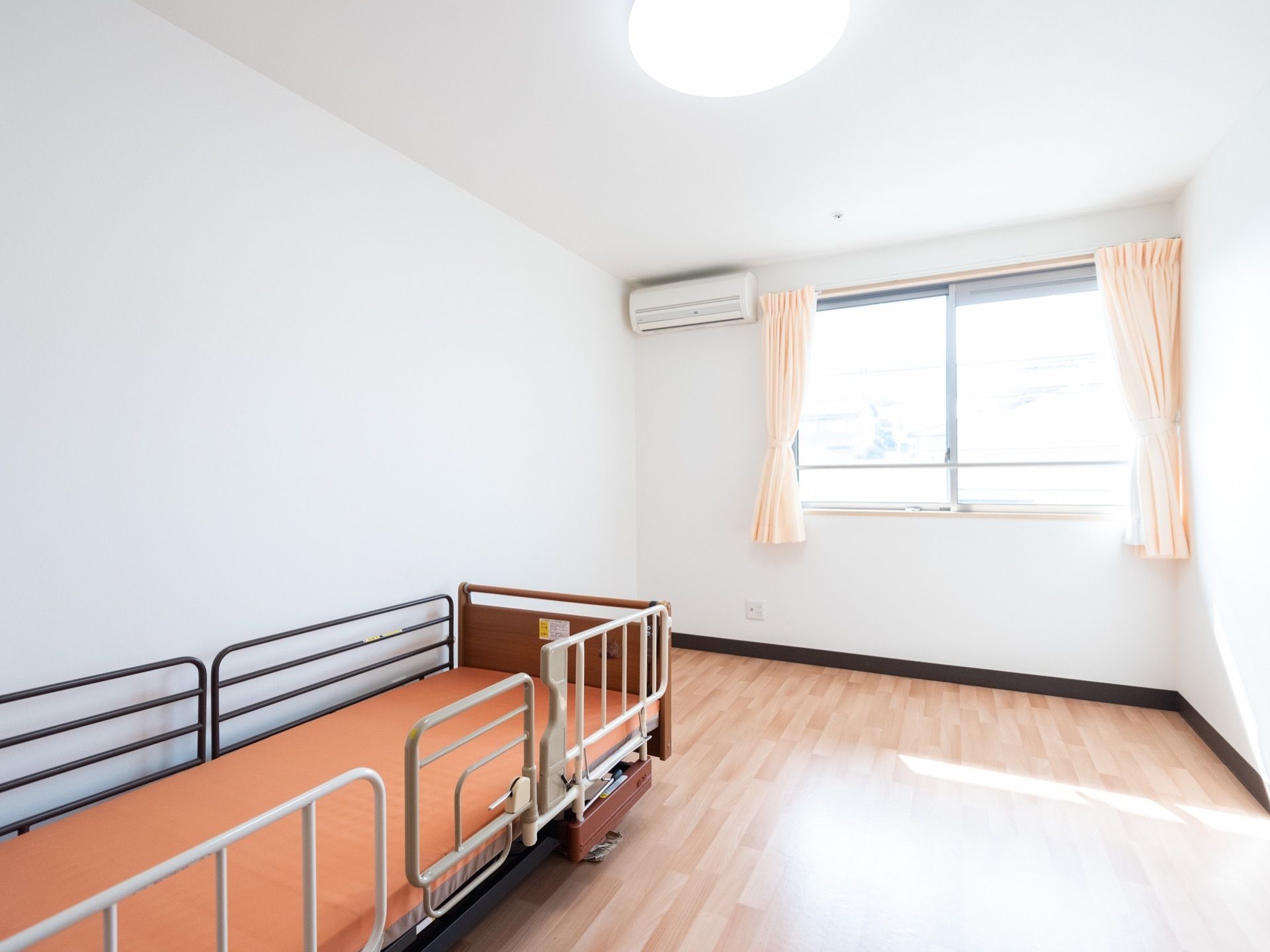 個室の部屋で、薄いピンクのカーテンが掛けられた腰掛け窓が設置されている。窓の左側にエアコンが取り付けられている。