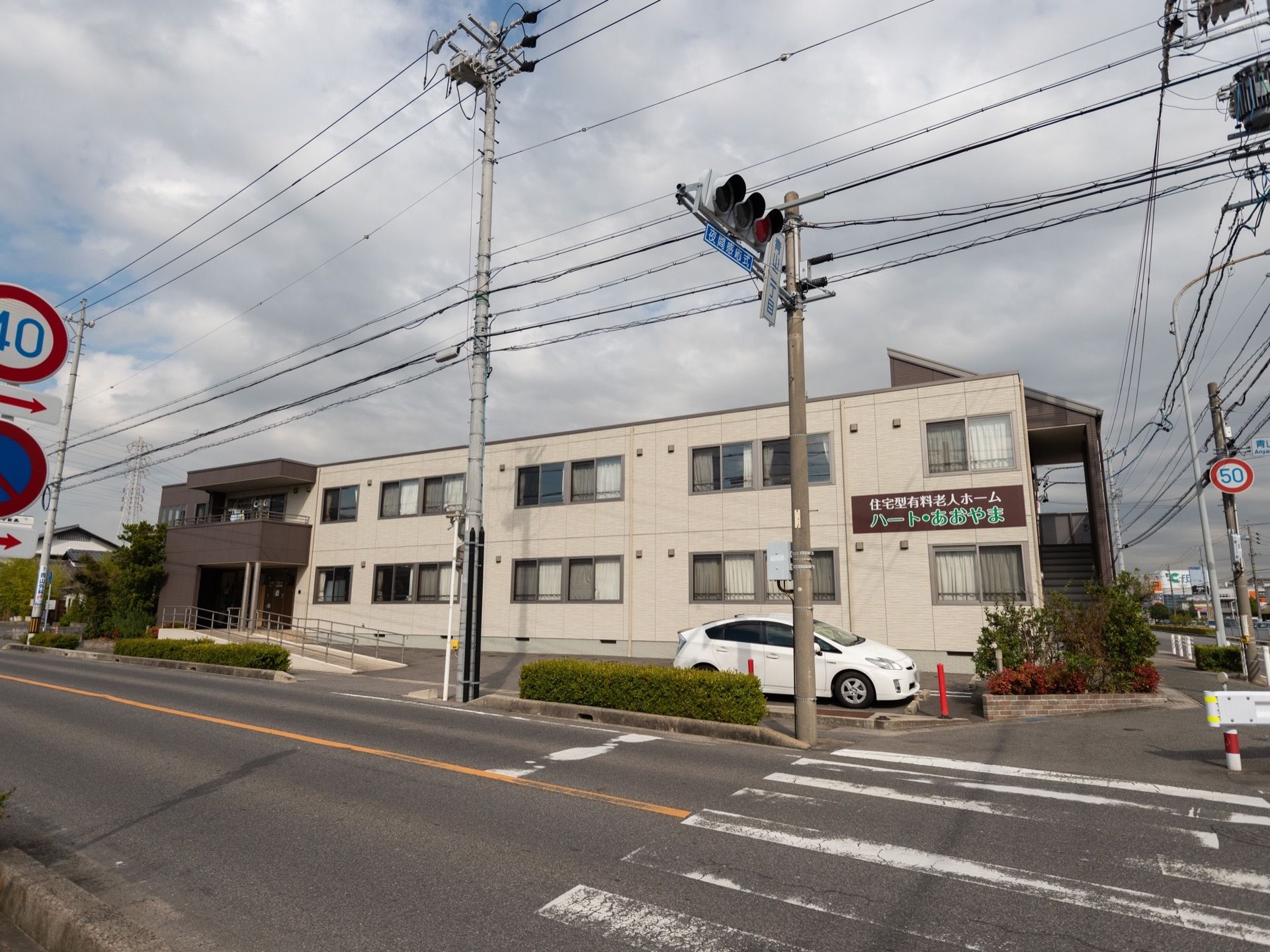2階建ての建物で、建物の前に白い車が1台停まっている。建物の右手前に花壇が作られており、樹木が植えられている。
