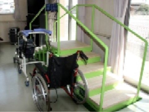 窓辺に鮮やかな緑色の歩行訓練階段が設置されている。その横には歩行器や車椅子が置かれている。スタッフのサポートを受けて歩行練習ができる。