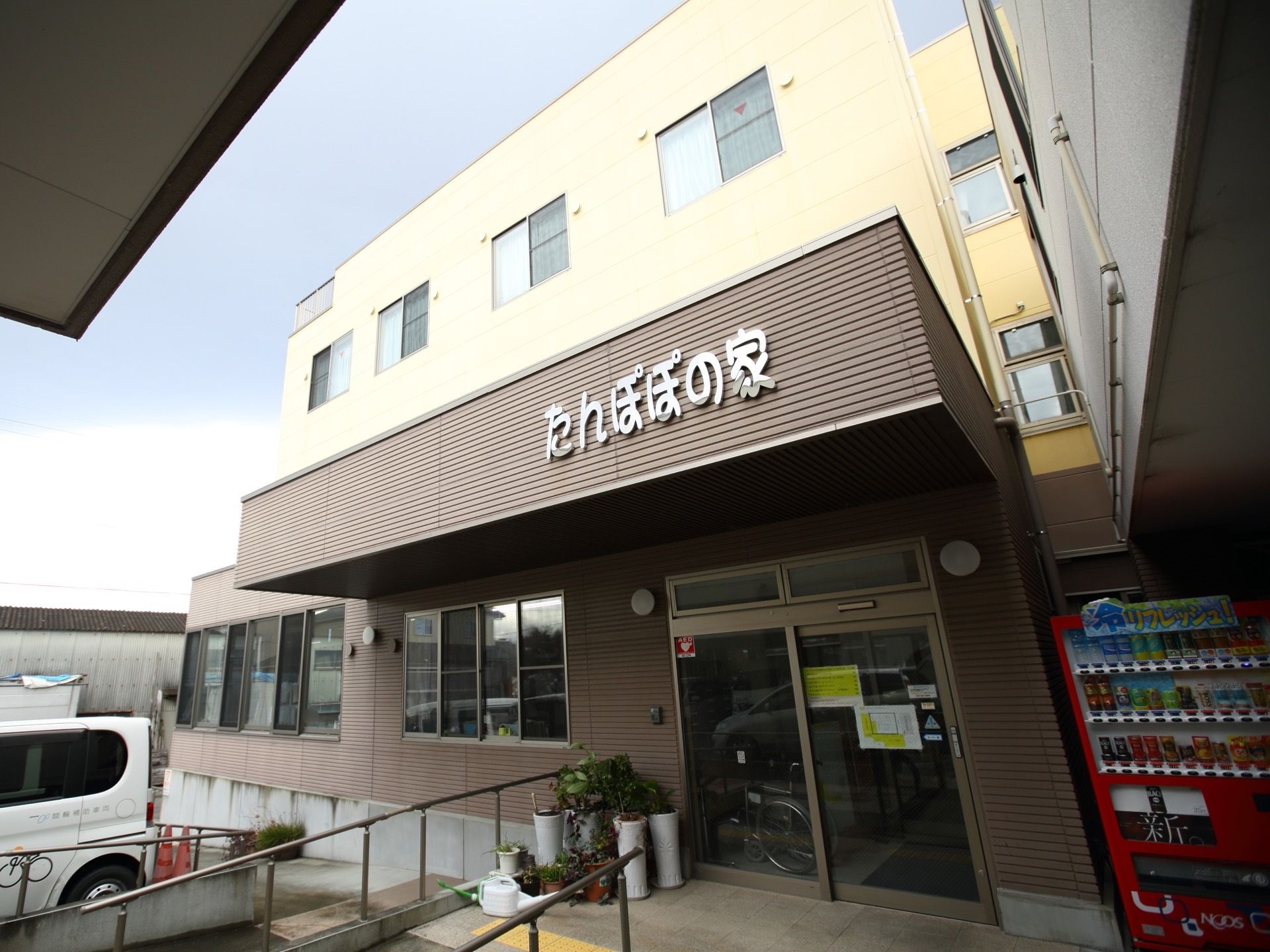 傾斜が緩やかな手すり付きのスロープが設置されている玄関である。床には、点字ブロックが設けられている。