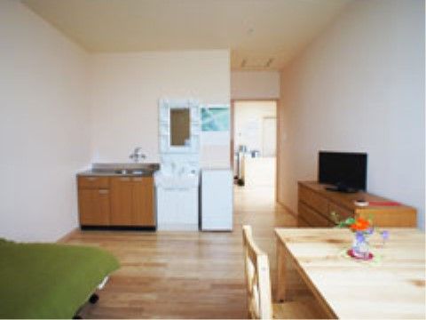 フローリングの床に白いクロスの壁である。茶色いチェストの上に小型の薄型テレビが置いてある。壁際に洗面台とシンクが設置されている。