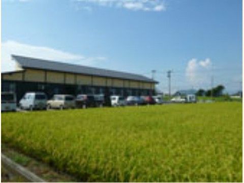 晴れ晴れとした空に緑が広がっている。緑が広がる近くには大きな屋根が付いた建物があり、9台ほどの車が止められている。