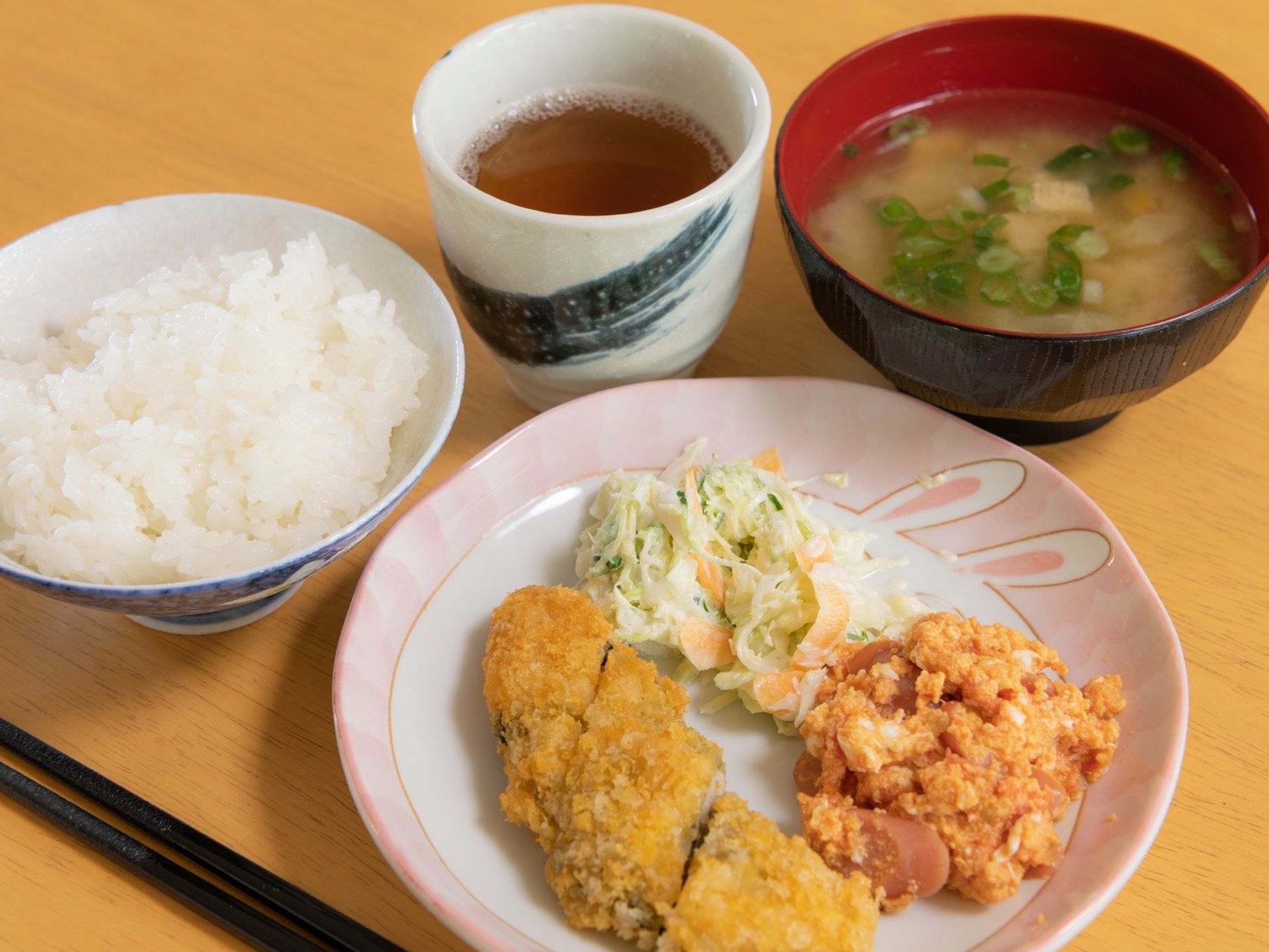 木質のテーブル上に食事が配膳されている。ご飯、味噌汁、お茶に加えて、数種類のフライと野菜が盛りつけられたイラスト付きのプレートがある。