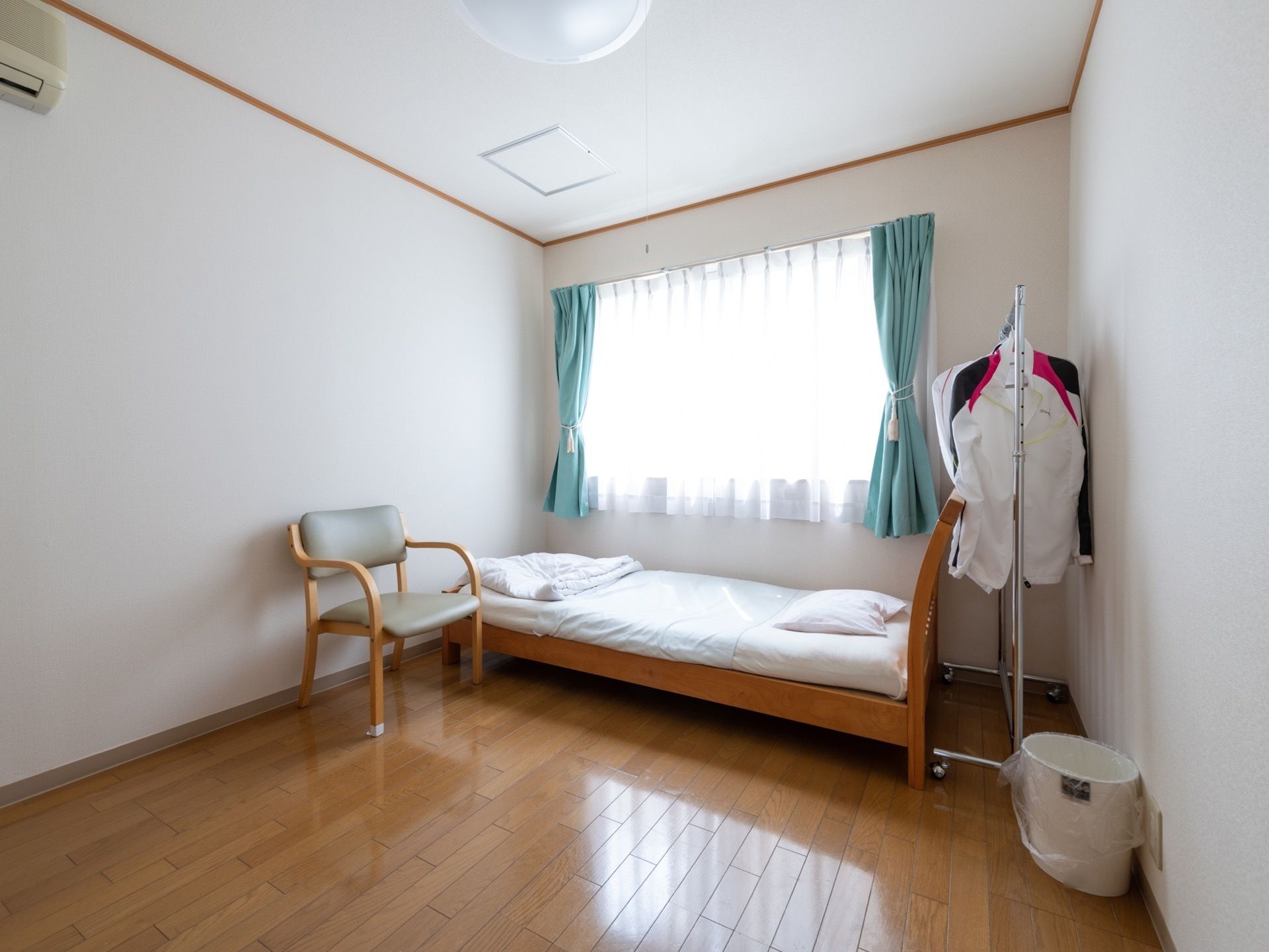 居室には大きな窓がある。きれいなフローリングの床で、ベッドや椅子、エアコン、衣装ハンガーが設置されている。