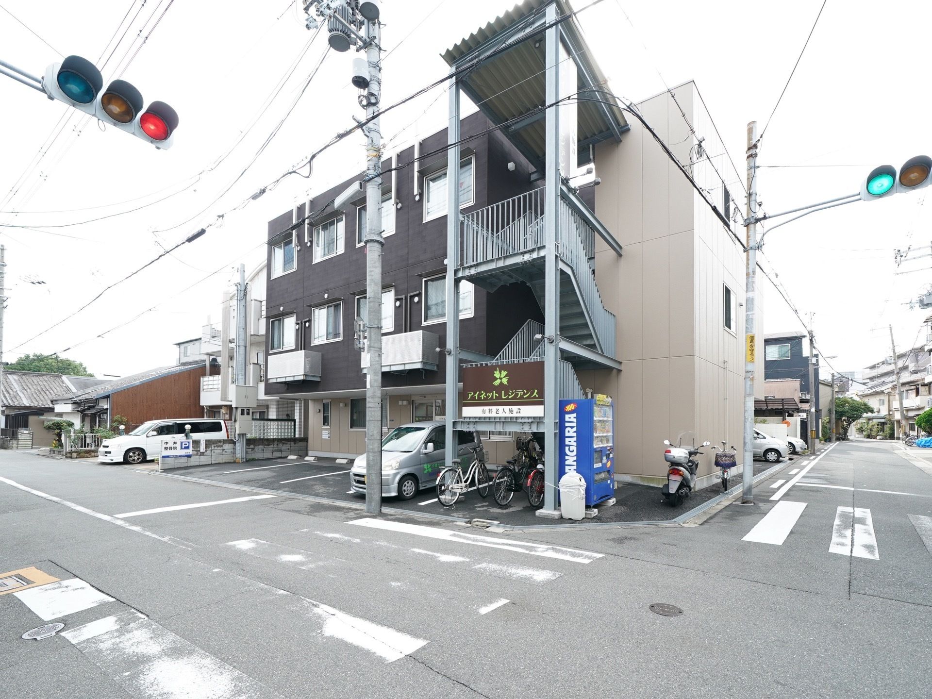 信号機のある交差点の角地に位置する３階建ての建物である。外階段が建物の脇に設置されており、２階の手すりの外側に施設名の看板が取りつけてある。