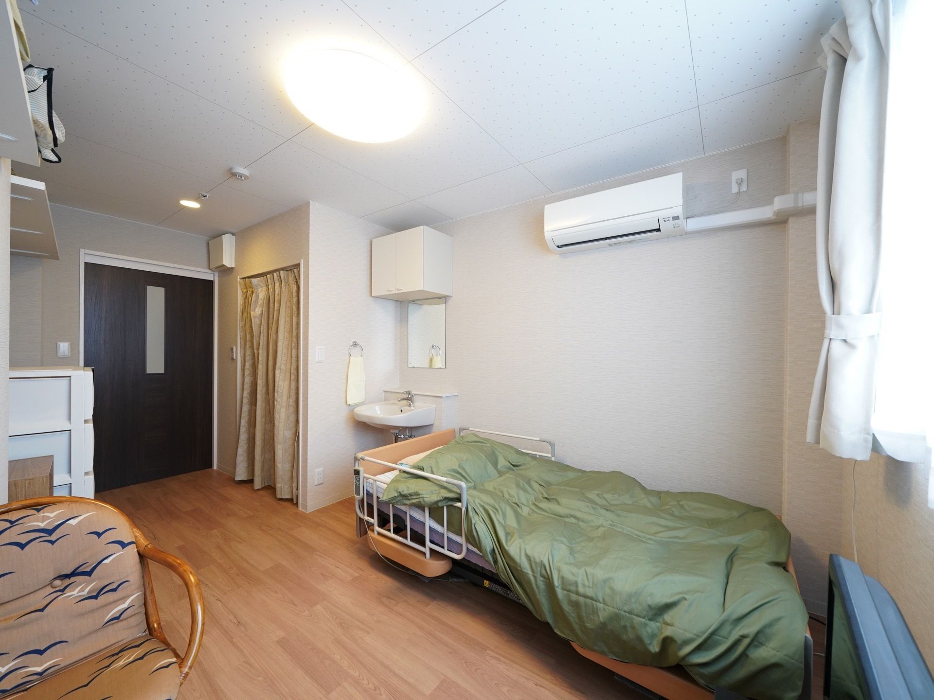 フローリングの床の部屋にベッドが置かれ、シーツが掛けられている。枕もとに洗面台が設置され、ベッド横の壁にはエアコンが取りつけられている。