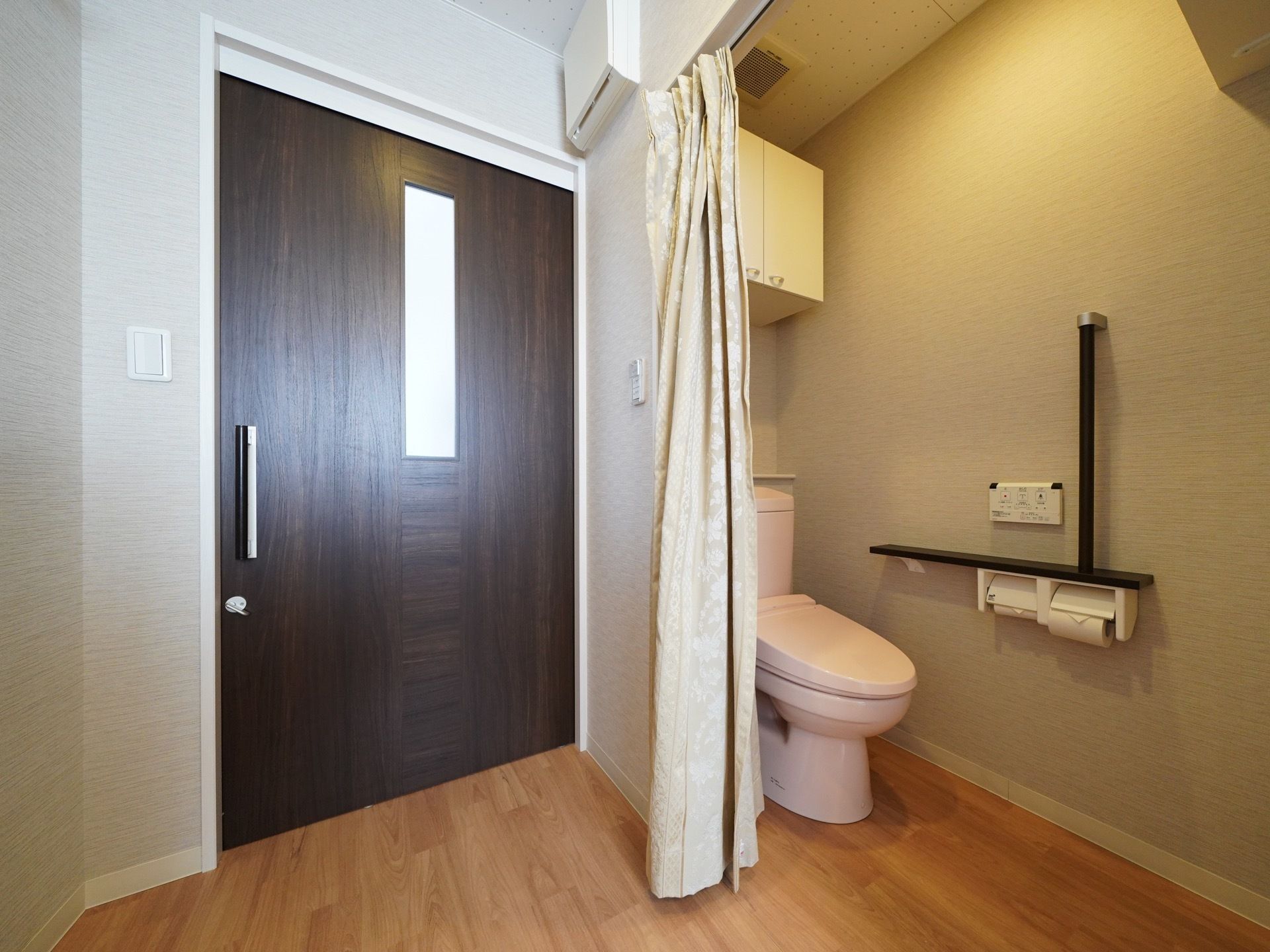 部屋の入口ドアの横にカーテンで仕切られたスペースがあり、洗浄機能付きのトイレが設置されている。便器脇の壁に操作盤と手すりが取りつけてある。