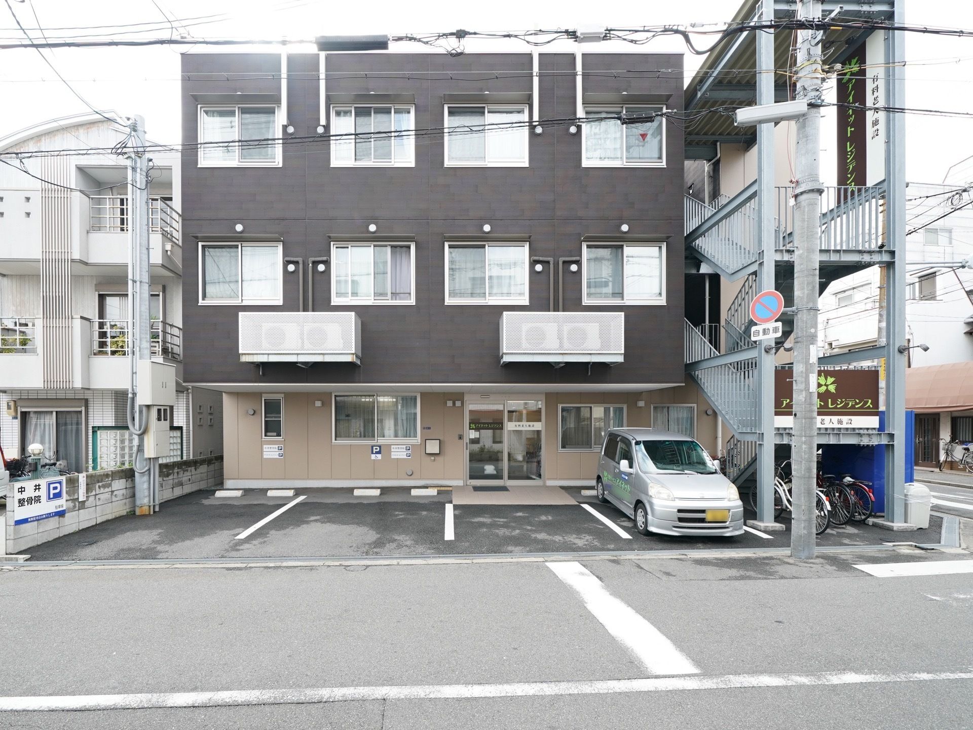 １階はクリーム色、２階と３階はダークグレーの外壁の３階建ての建物である。建物の右側に外階段が設置されている。施設の前に駐車スペースがある。