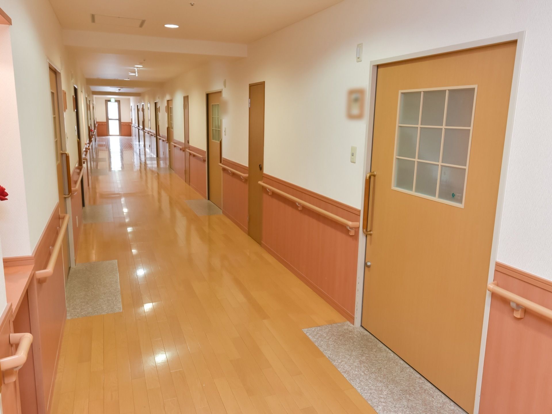 広いスペースの廊下である。両サイドに並んでいる居室ドアの横には、チャイムが設置されている。壁には、手すりが備わっている。