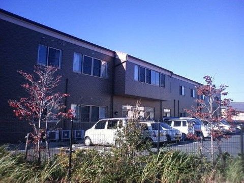 駐車場のすぐ前に、施設の建物が建てられている。茶色い壁には窓が多くあり、地面にエアコンの室外機が並べて設置されている。