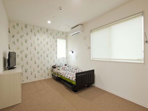 壁はボタニカルな模様である。介護用ベッドが置いてあり、反対の壁には衣装ケースが用意され、その上にはテレビがある。