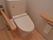 トイレの個室内。木材の床で、壁は白色をしている。トイレの操作ボタンが、手元についている。また右手に手すりが設置されている。