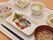 食事メニューの一例。赤飯、芋と油揚げの味噌汁、魚料理に加え、卯の花と野菜の煮浸しがある。お茶も添えられている。