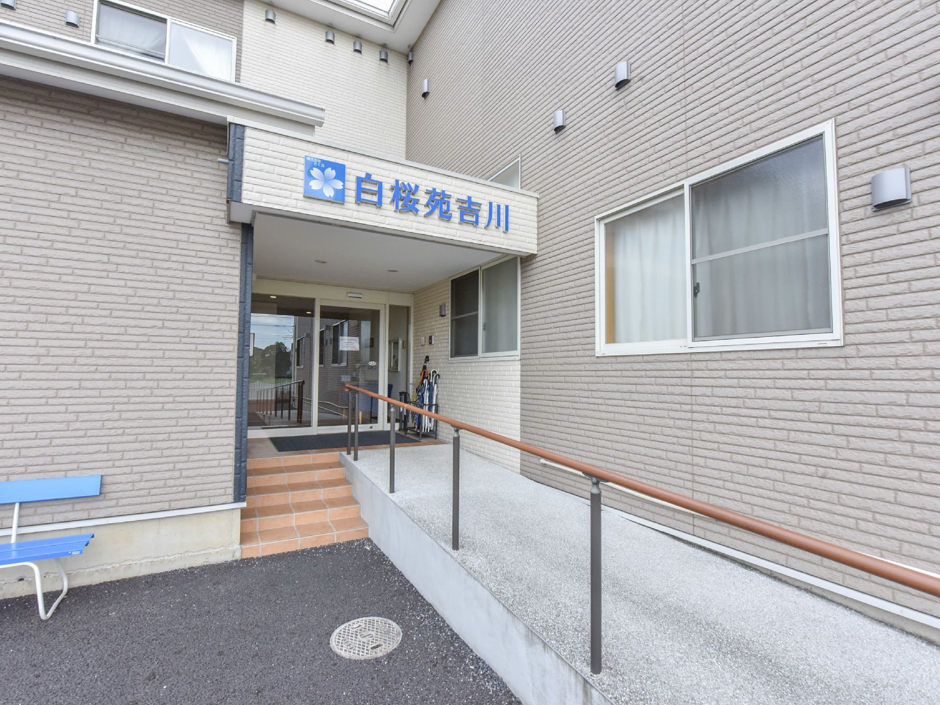 施設の入口へは、階段またはスロープを利用できる。建物は、レンガ造り風の壁である。建物の前に、ベンチが置かれている。