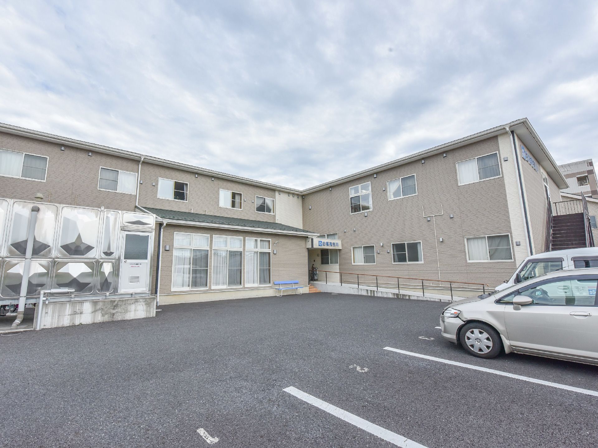 施設の建物の前には、駐車スペースがあり、番号が振られている。施設の向かって左手には、銀色の建物が見える。