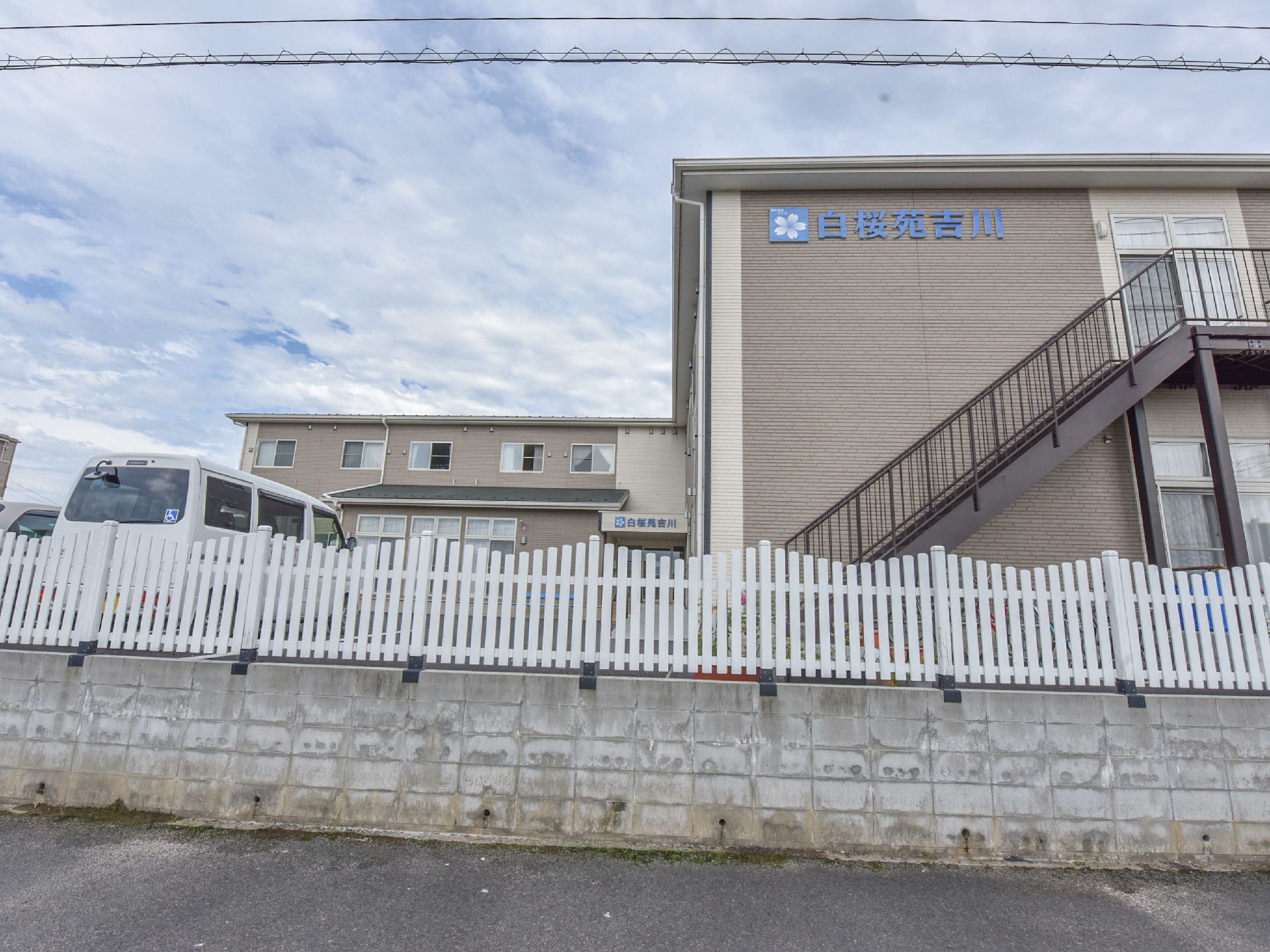 施設は、ブロック塀が重なっている高い位置にあり、その上には、白い板が組まれたフェンスがある。施設の外に、外階段がある。