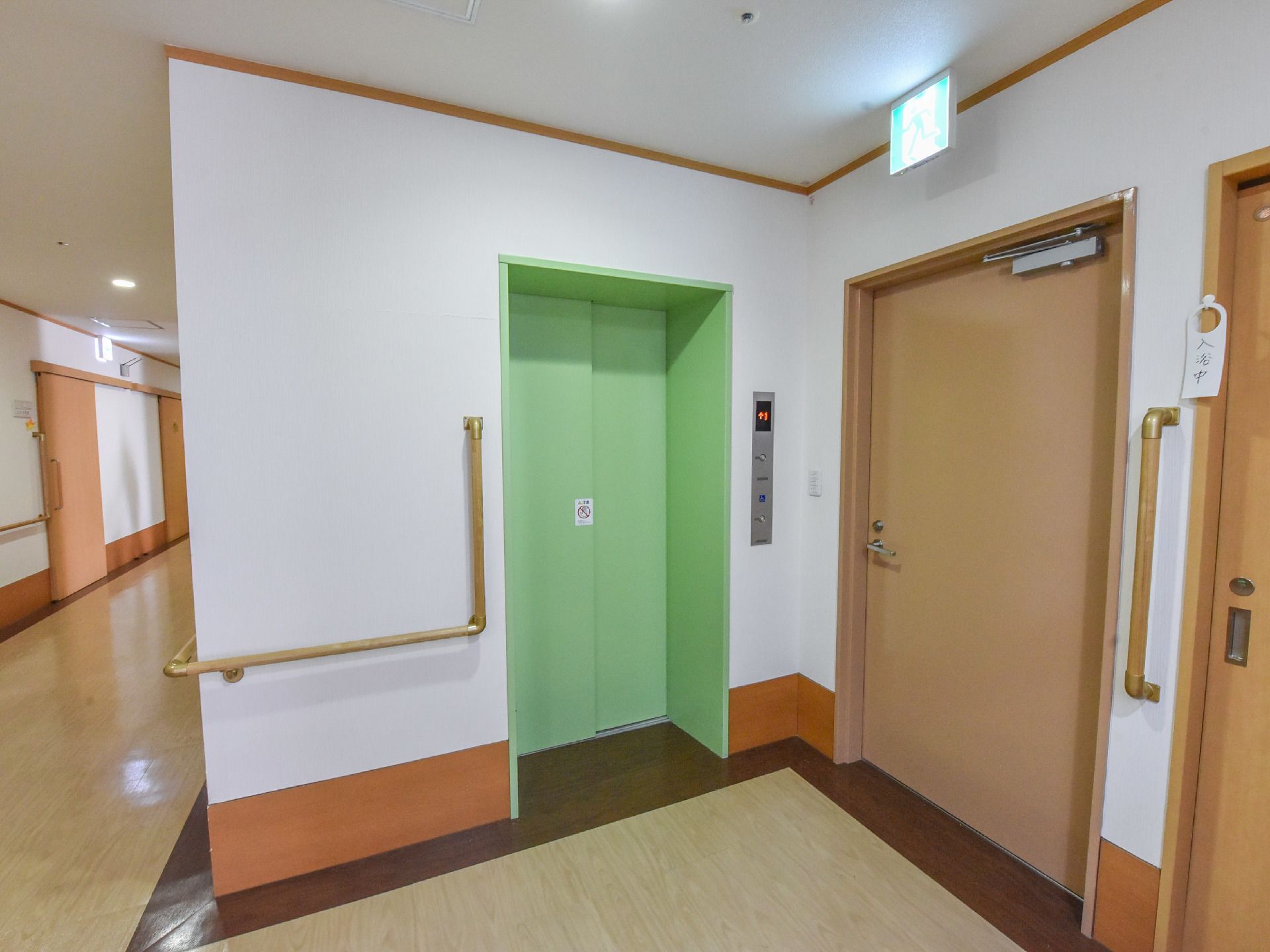 施設内に、エレベーターが完備されている。ドアは黄緑色で、周りの壁は白色である。右手に茶色いドアがある。