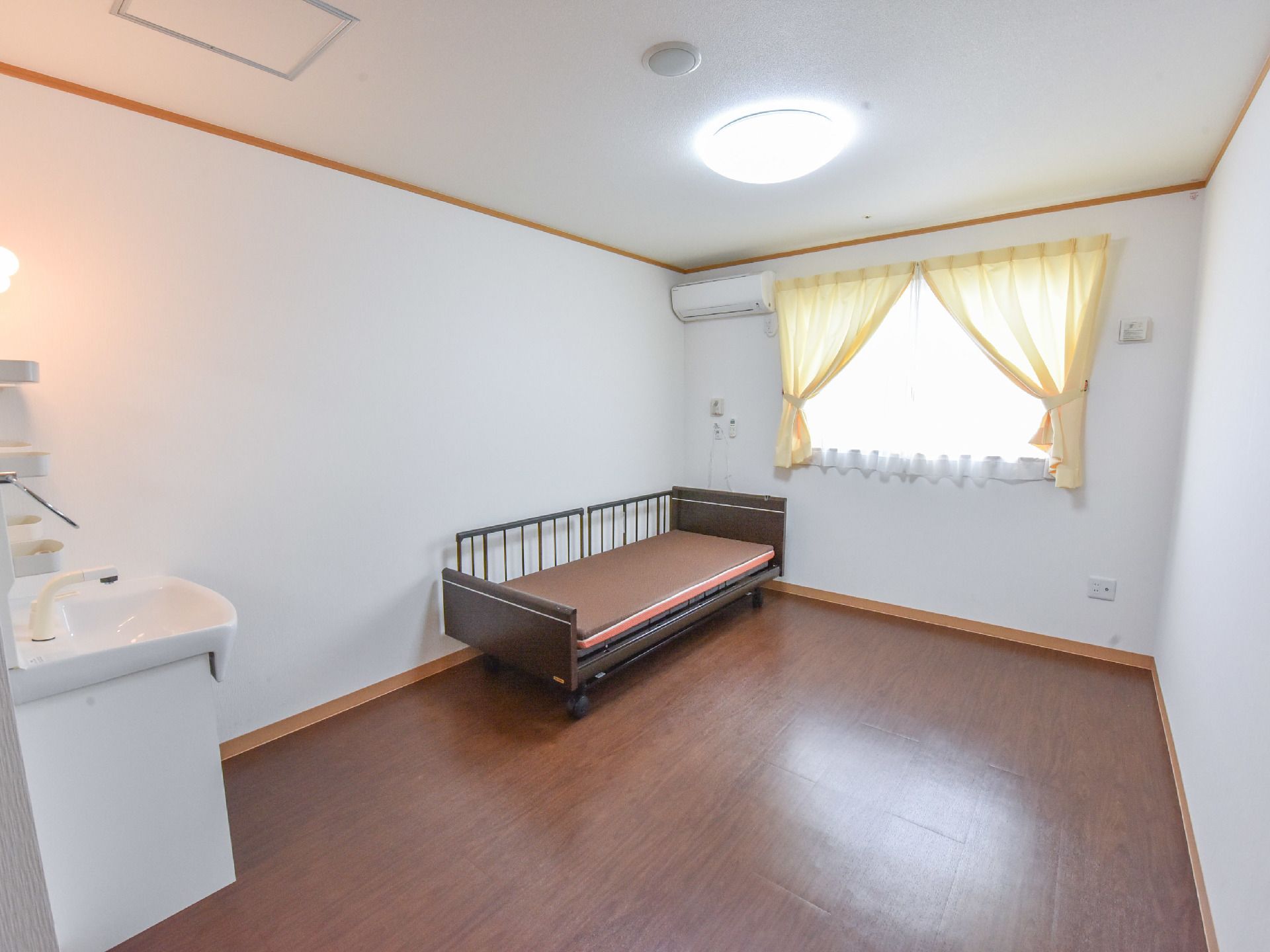 居室の壁と天井は白色で、床はこげ茶色のフローリングである。ベッドがあり、その足元に洗面台が設置されている。