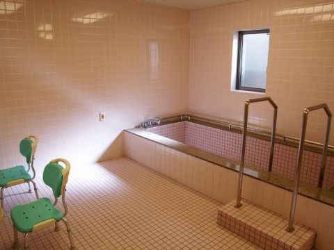 ピンクのタイル張りの浴室。洗い場には高めの椅子が設置されており、かがむことなく安全に座ることができる。浴槽の周りには手すりがある。