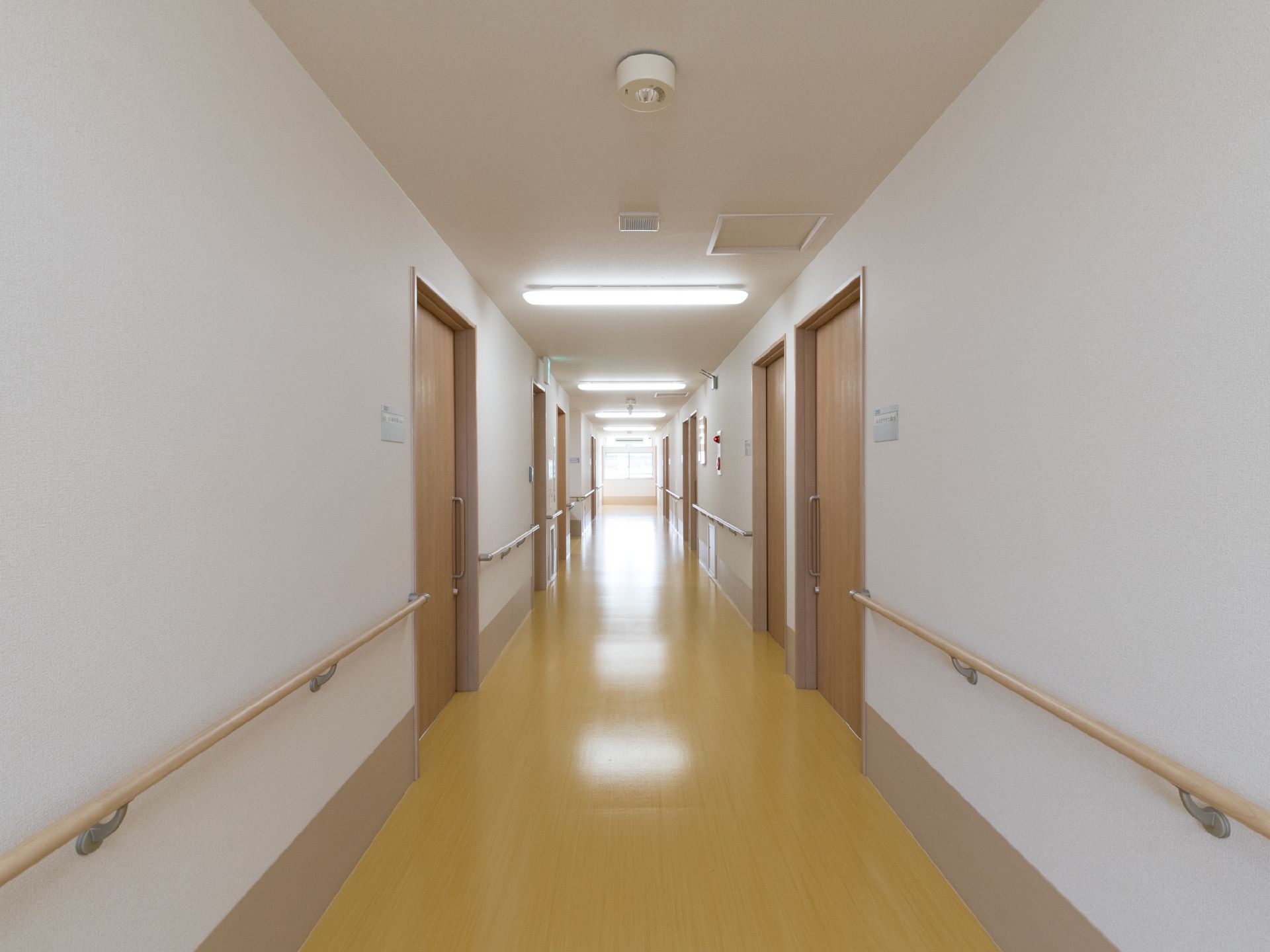 薄い茶色のフローリングの床の廊下がまっすぐ続いている。壁の両側には手すりが設置されている。部屋の扉もある。