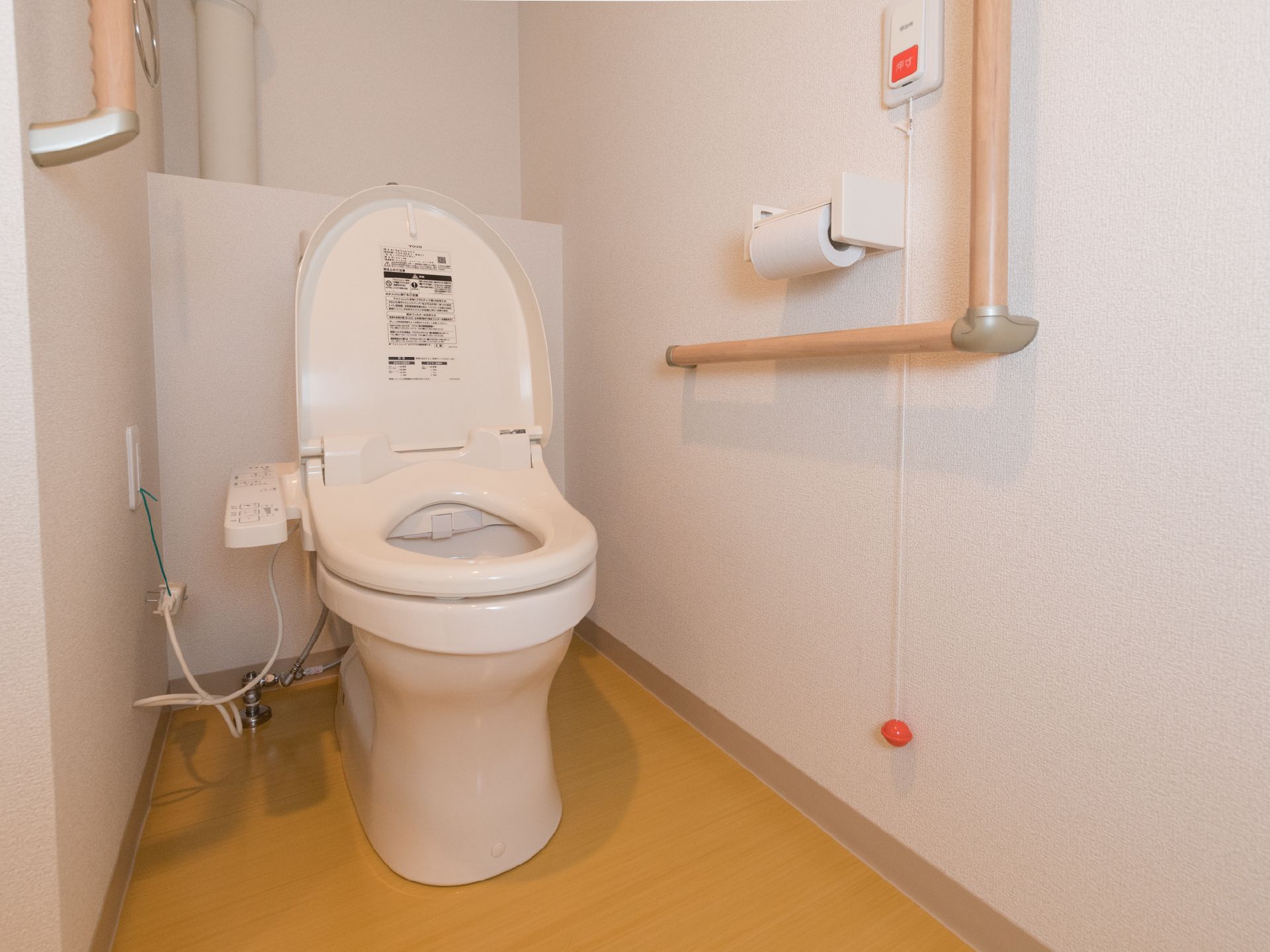 洋式便器が設置されたトイレがある。壁側には形状が違う手すりが取り付けられている。トイレットペーパーフォルダー側には呼び出しボタンもある。