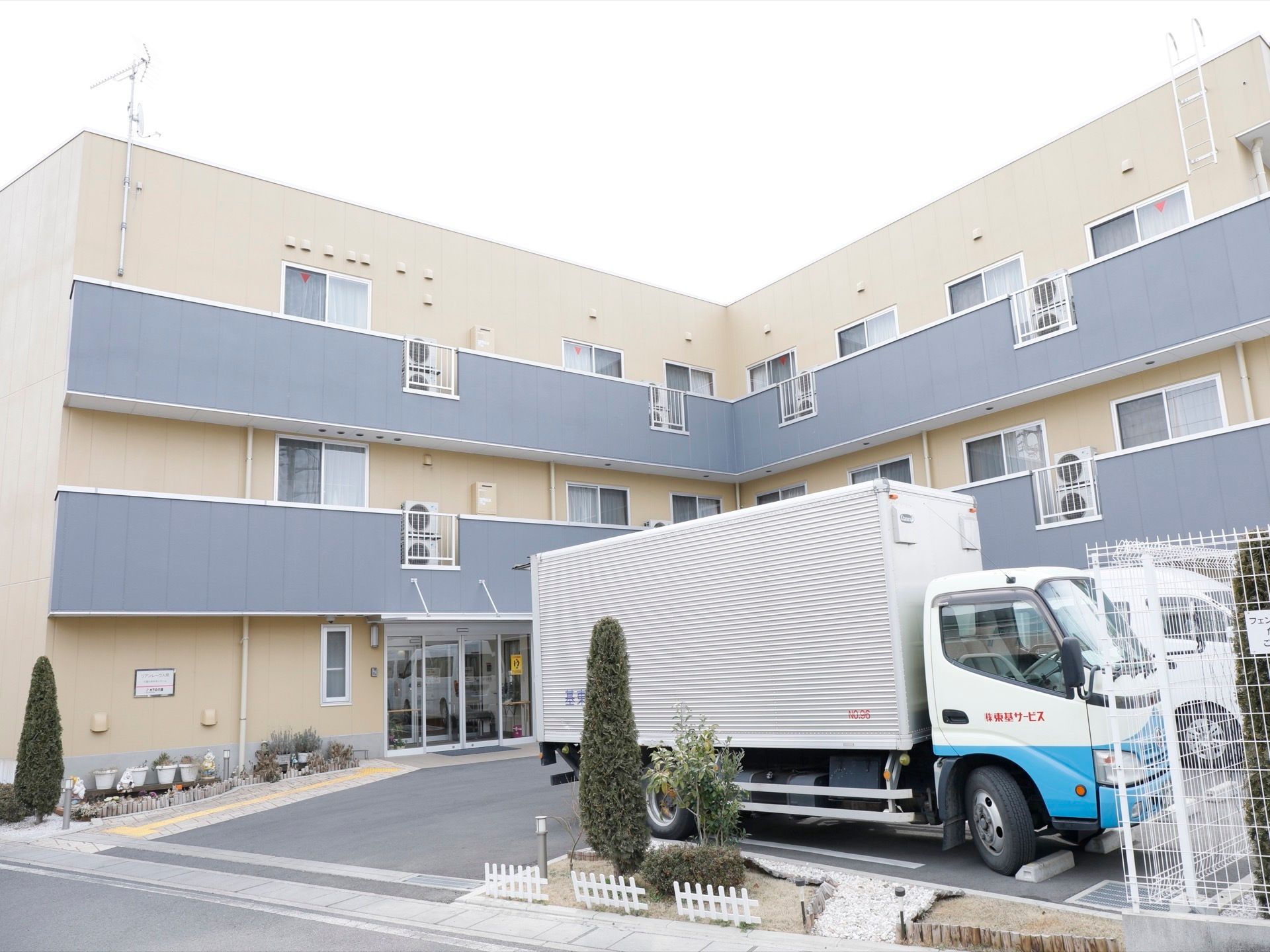 建物全体はベージュ色で、バルコニー部分だけペールグレーでデザインされた落ち着いた雰囲気の外観である。駐車場には軽トラックが置いてある。
