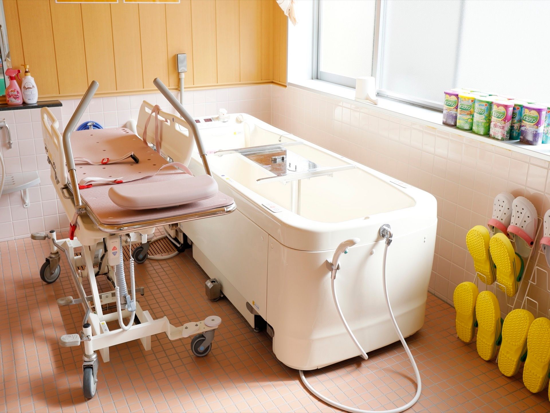 寝浴用の介護機器が、壁際に設置されている。右端には黄色いバスブーツが置いてある。その上の窓際に、たくさんの入浴剤が並んでいる。