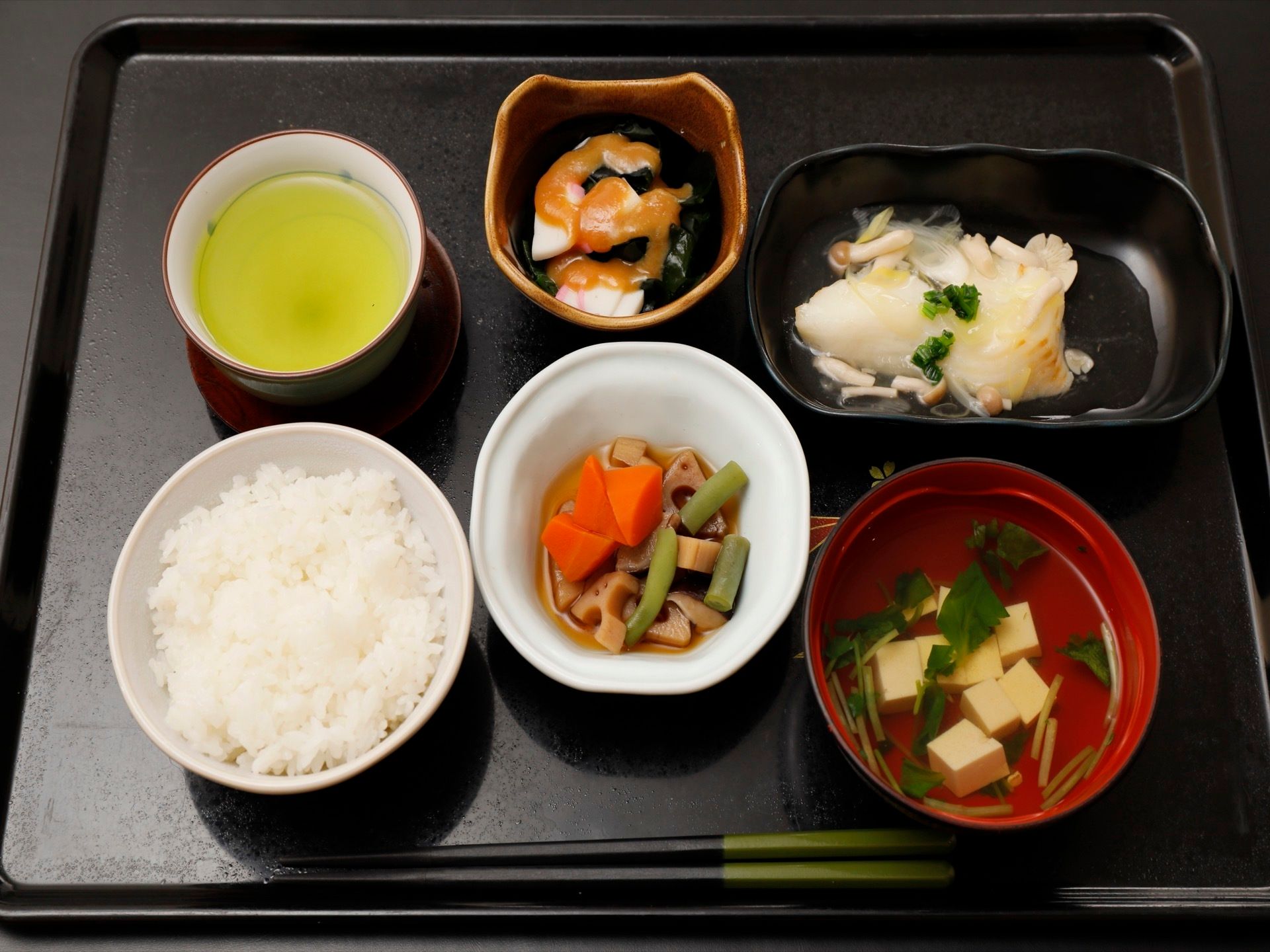 黒いトレーに、白米、豆腐のすまし汁、ニンジンやレンコンが入った煮物、白身魚のキノコあんかけ、小鉢に入ったワカメなどが並んでいる。