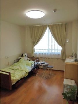 介護ベッドや机の置かれた居室は、フローリングの床と白色の壁である。天井にはスプリンクラーが設置されている。