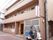 ３階建ての落ち着いた色合いの外壁の建物。各部屋にはベランダが設置されており、施設前にバイクが置かれている。