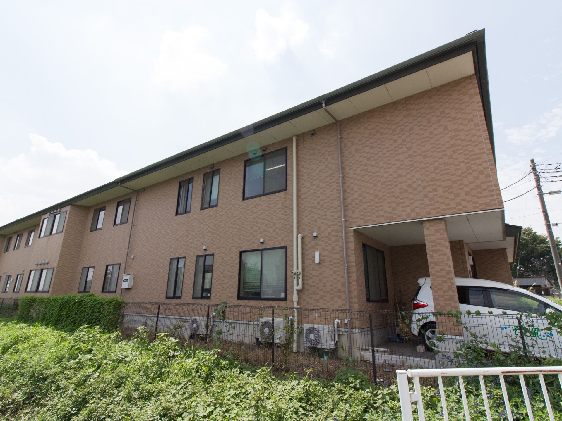 ブラウンを基調とした落ち着いた色合いの２階建ての建物。壁沿いには、エアコンの室外機が複数置かれている。
