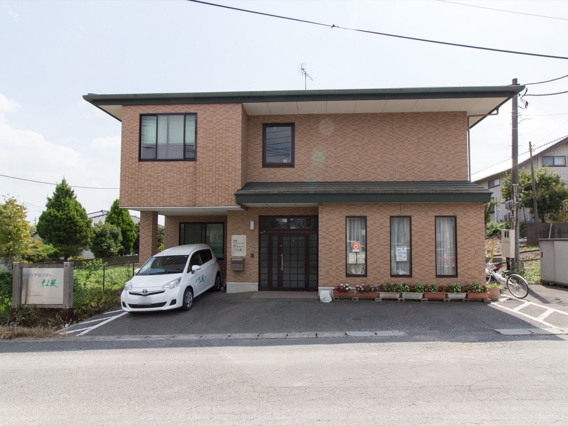 閑静な住宅街の中にあるグループホームである。アスファルトで舗装された駐車場には、白い施設専用車が停められている。