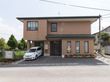 閑静な住宅街の中にあるグループホームである。アスファルトで舗装された駐車場には、白い施設専用車が停められている。