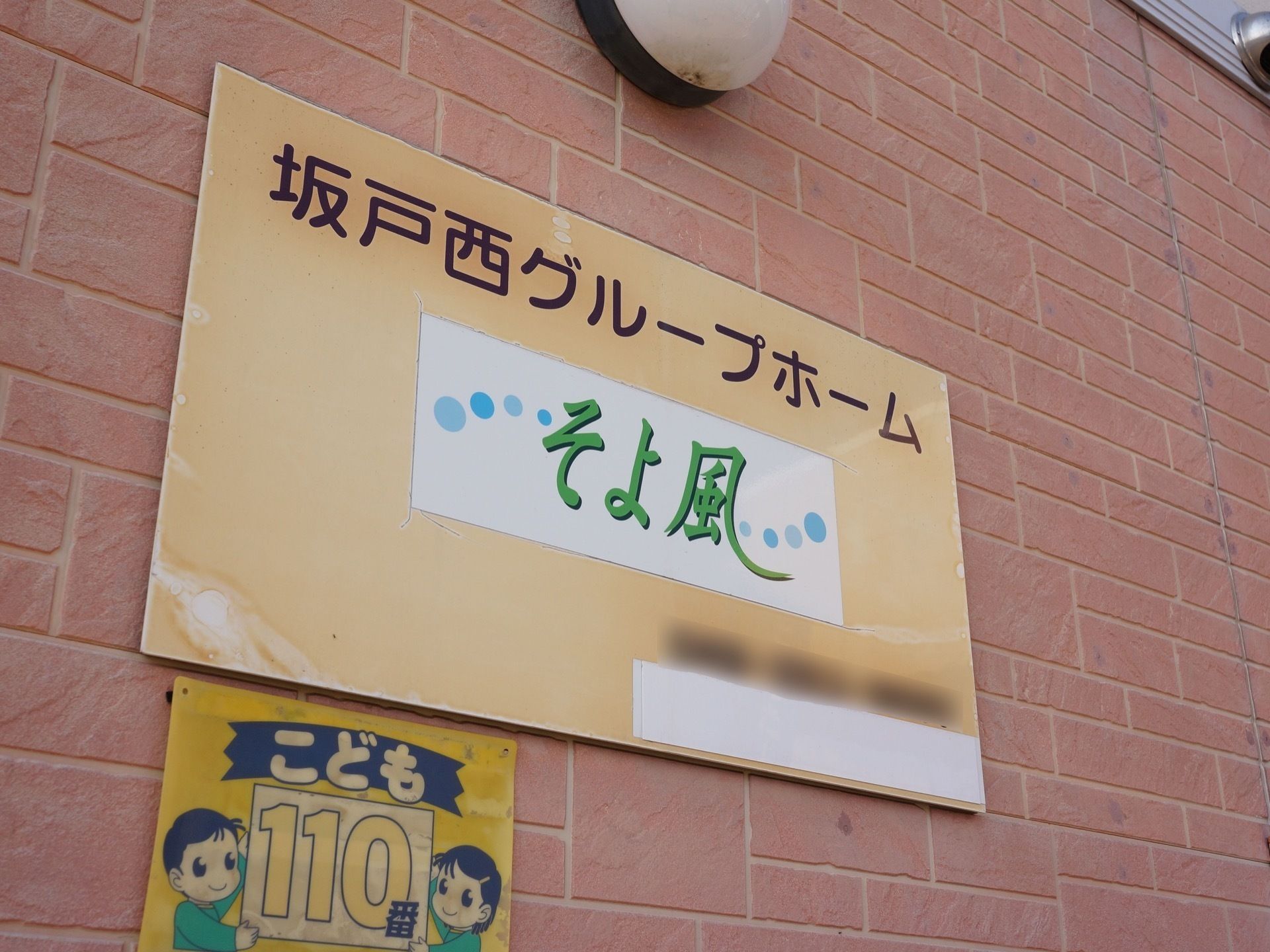 施設の外壁に看板が設置されている。看板の上に丸い照明がついている。また、下には子供の緊急時に対応可能な場所を示す防犯プレートがある。