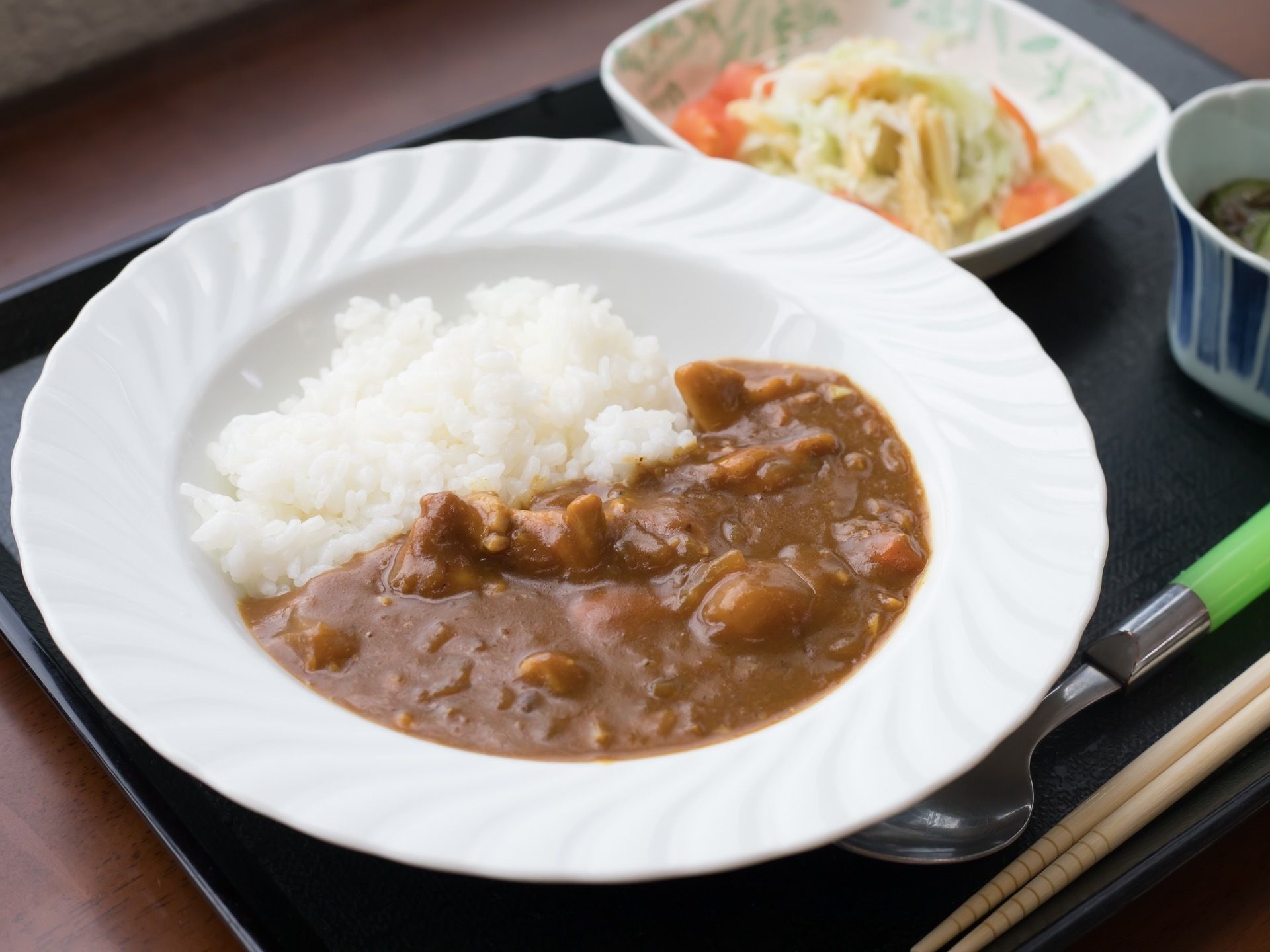 テーブルの上に置かれた黒いトレイには、カレーや野菜のおかず、茶碗蒸しが並べられている。箸とスプーンが用意されている。