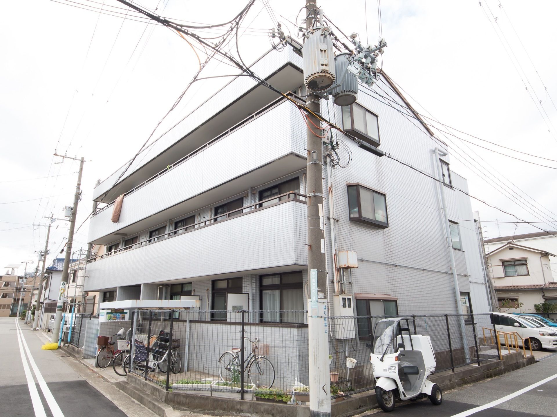 道路の角地に建つ、３階建ての建物である。玄関のある面に駐車スペースがあり、もう一方の面に自転車置き場がある。