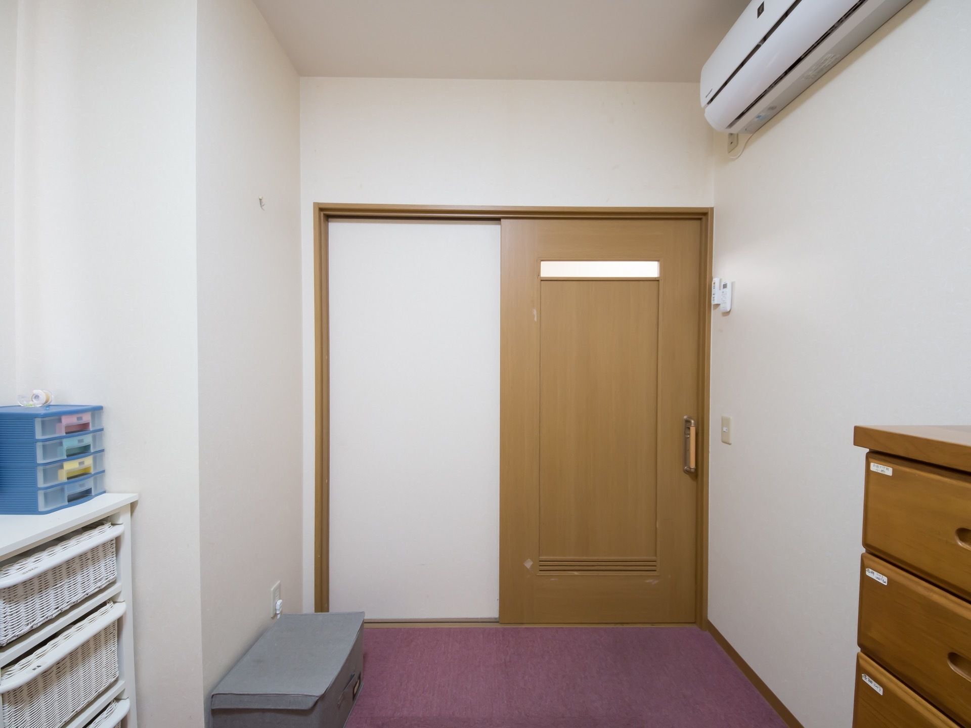 入口は引き戸で、白い壁に落ち着いた濃いピンク色のカーペット敷きの部屋である。収納棚やエアコンが設置されている。