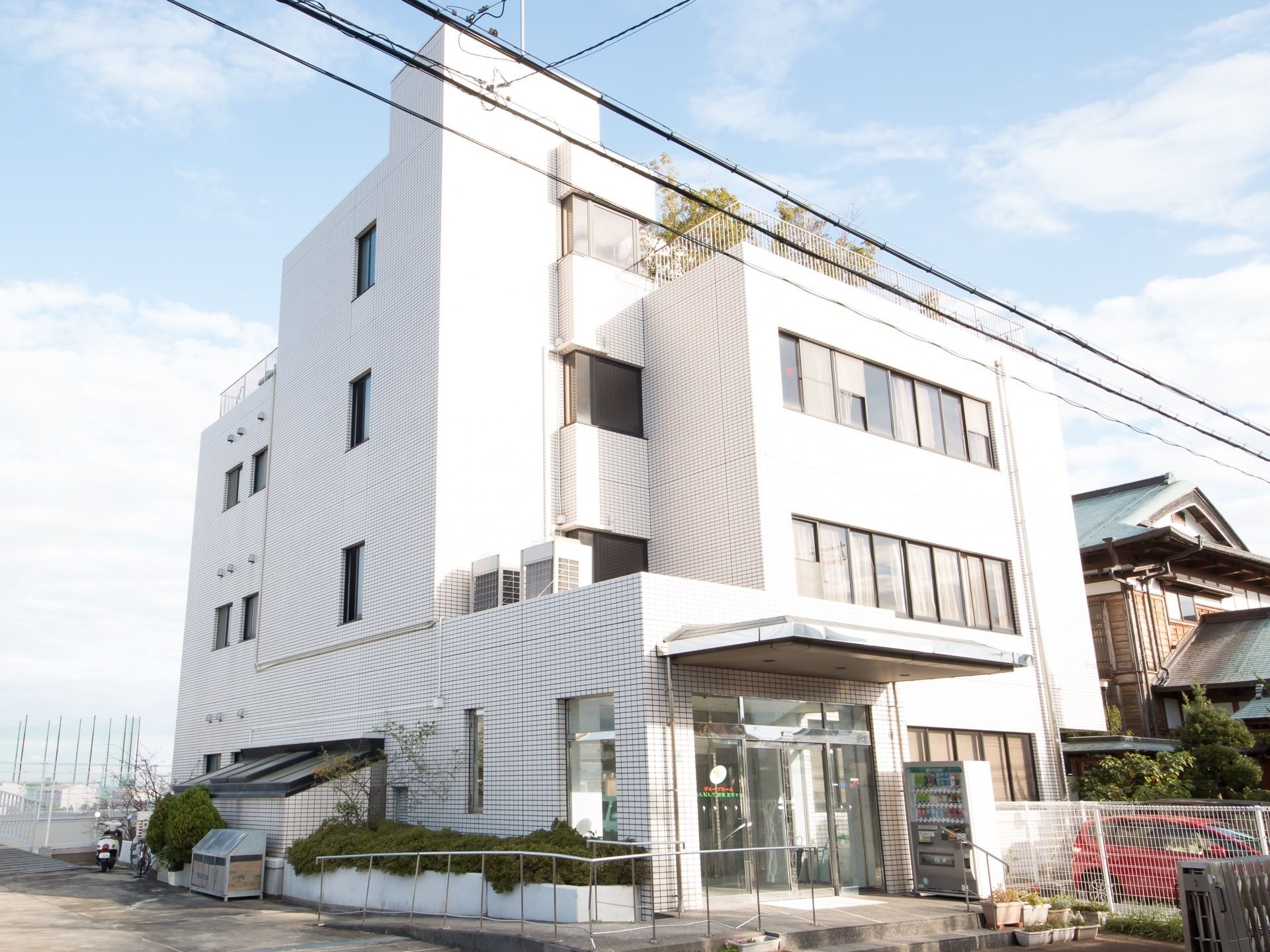 タイル張りの建物で、玄関上に大きなひさしがついている。自動販売機とスロープが設置されている。屋上に木があるのが見える。