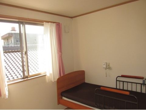 ワンルームの居室に、茶色のマットが敷かれたベッドが置かれている。横の壁にはナースコールが取り付けられている。