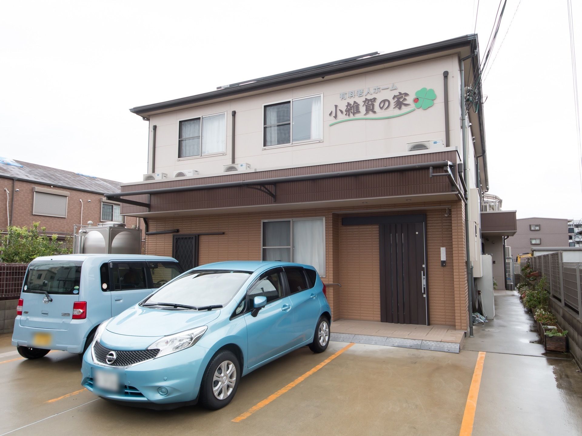 ２階建ての施設の前に２台の車が停められている。玄関には施設を感じさせない一般住宅と同じようなドアを採用している。