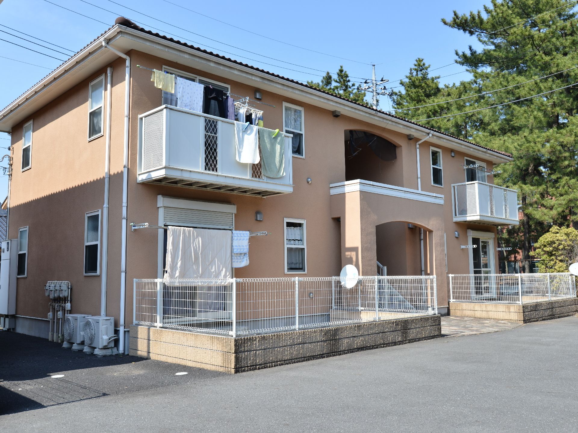 集合住宅のようなつくりで1階に2つの庭と2階に2つのベランダがみえる。建物右側には豊富な木々をみてとれる。