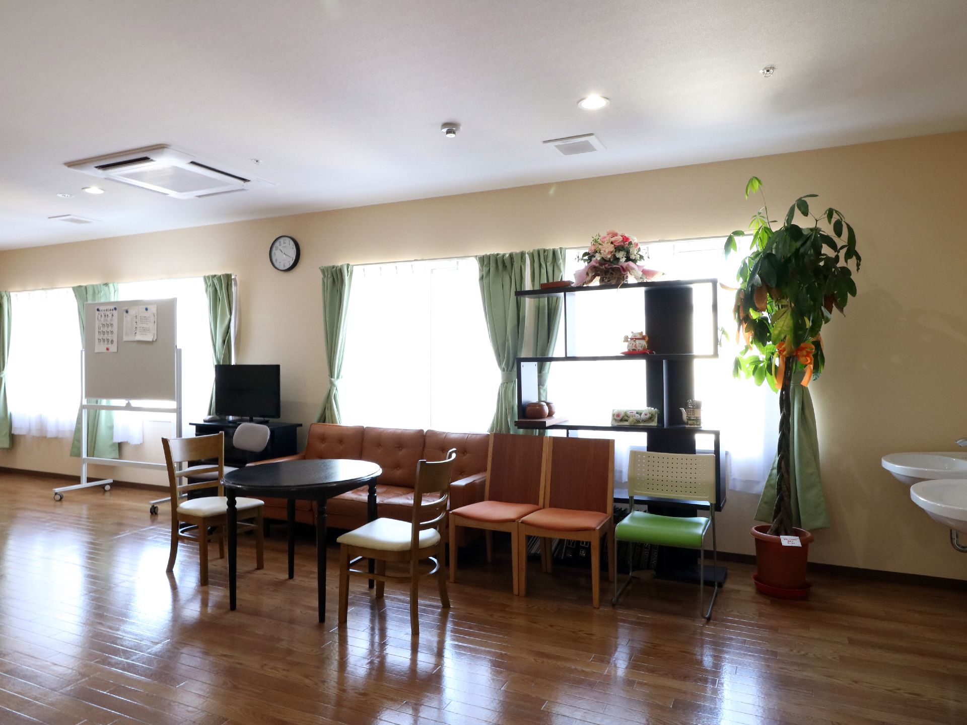 談話室は広く長い空間でゆとりがある。掲示板や丸テーブル、ソファやテレビなどのほか観葉植物や置物なども飾っている。