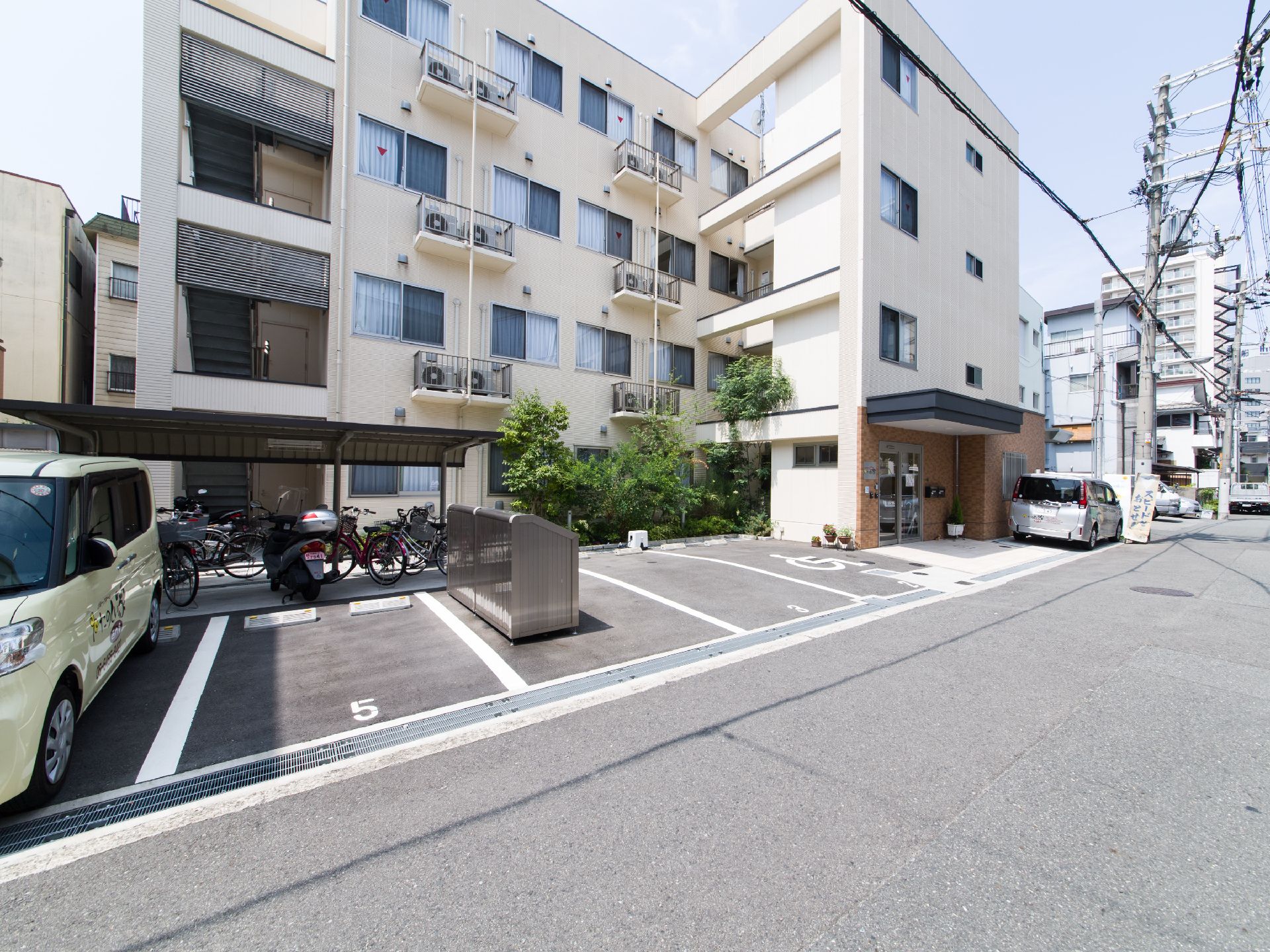 正面には4階建ての建物が建っている。その手前には駐輪場があり、そこのさらに手前には駐車場がある。建物は道路に面している。