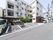 正面には4階建ての建物が建っている。その手前には駐輪場があり、そこのさらに手前には駐車場がある。建物は道路に面している。