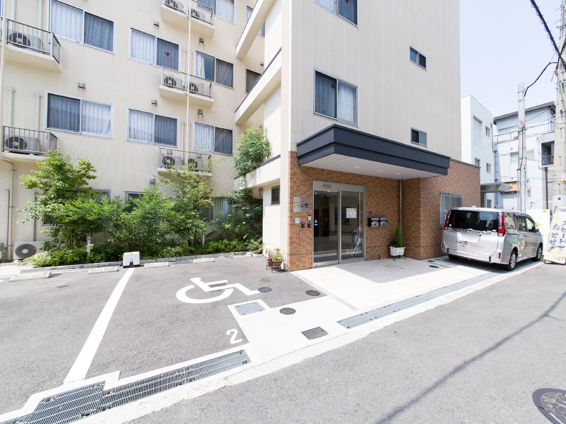 建物が正面に立っている。右側には玄関があり、そばには車が1台止められている。外壁は白色に塗られている。